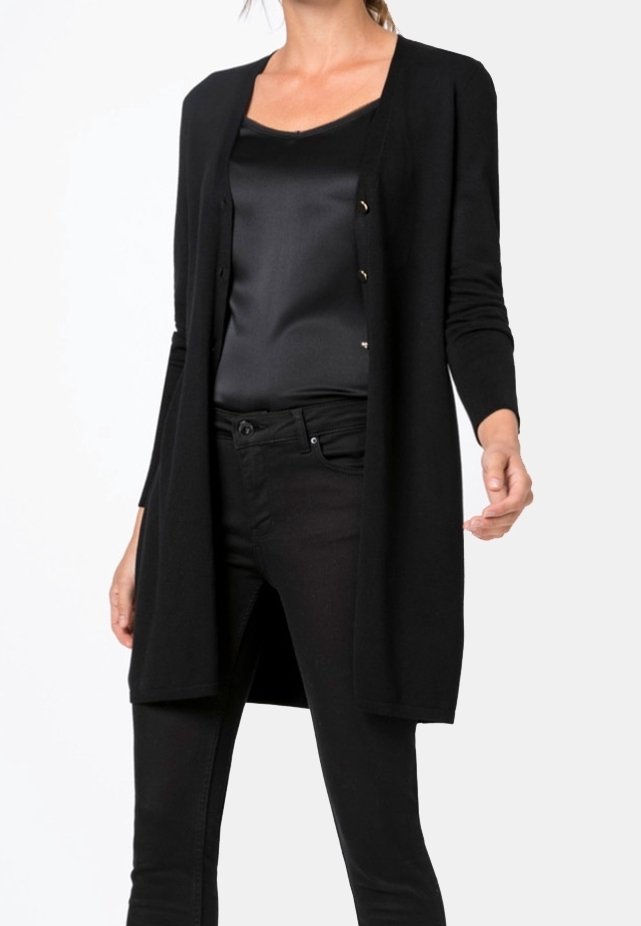 Femme portant un long cardigan noir par-dessus un caraco en satin noir et un pantalon noir, debout devant un fond blanc.