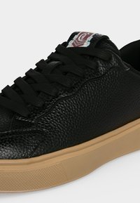 Μαύρο sneaker από υφή δέρματος με μαύρα κορδόνια και ταμπά καουτσούκ σόλα, με κόκκινο και λευκό λογότυπο στη γλώσσα.