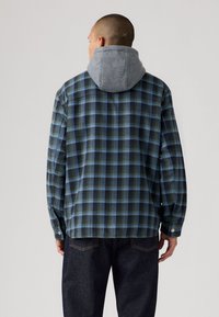 Plaid flannel shirt in blue and green, featuring a grey hoodie attached at the collar. Long sleeves with button cuffs, straight hem.