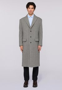 Dlouhý šedý vlněný kabát s klopy, zap�ínání na jeden knoflík, boční kapsy a rovný střih, doplněný o světle modrou košili a tmavé kalhoty.