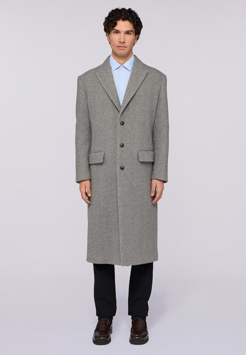 Dlouhý šedý vlněný kabát s klopy, zapínání na jeden knoflík, boční kapsy a rovný střih, doplněný o světle modrou košili a tmavé kalhoty.