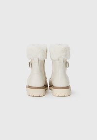 Bottes de cheville couleur crème avec un revers en fausse fourrure, fermeture éclair arrière et semelle en caoutchouc beige clair. Présente un accent de boucle sur le côté.