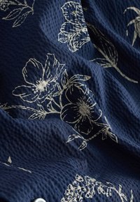 Marineblauer Stoff mit einem strukturierten Muster, das weiße Blumenmotive zeigt. Die Textur wirkt erhaben und gewebt, was das visuelle Interesse erhöht.