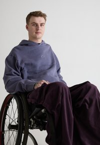 Hanorac purpuriu cu buzunar frontal și logo, asortat cu pantaloni largi de culoare purpurie închisă. Modelul este așezat într-un scaun cu rotile, pe un fundal neutru.