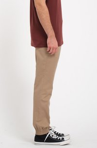 Pantalons beige fuselés avec des poignets élastiques, associés à des baskets noires avec des semelles et des lacets blancs. Une chemise bordeaux foncé est visible.