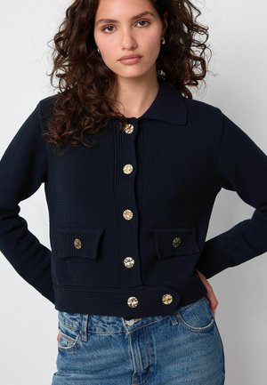 Navy knitted cardigan with a collar, gold decorative buttons, and two front pockets. Features ribbed cuffs and hem.