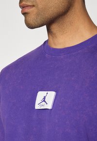Lilla bomulds T-shirt med en tekstureret overflade, der har et hvidt rektangulært mærke med et lilla Jumpman-logo og signatur.