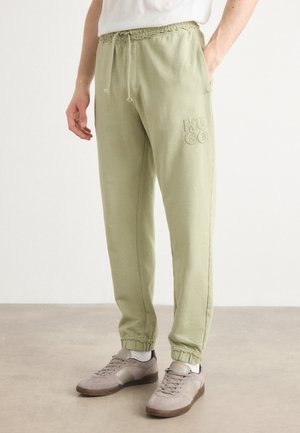 Mann trägt hellgrüne Jogginghosen mit elastischem Bund und dem Schriftzug „HUGO“ am Oberschenkel sowie beige Sneakers mit weißen Socken auf neutralem Bodenhintergrund.