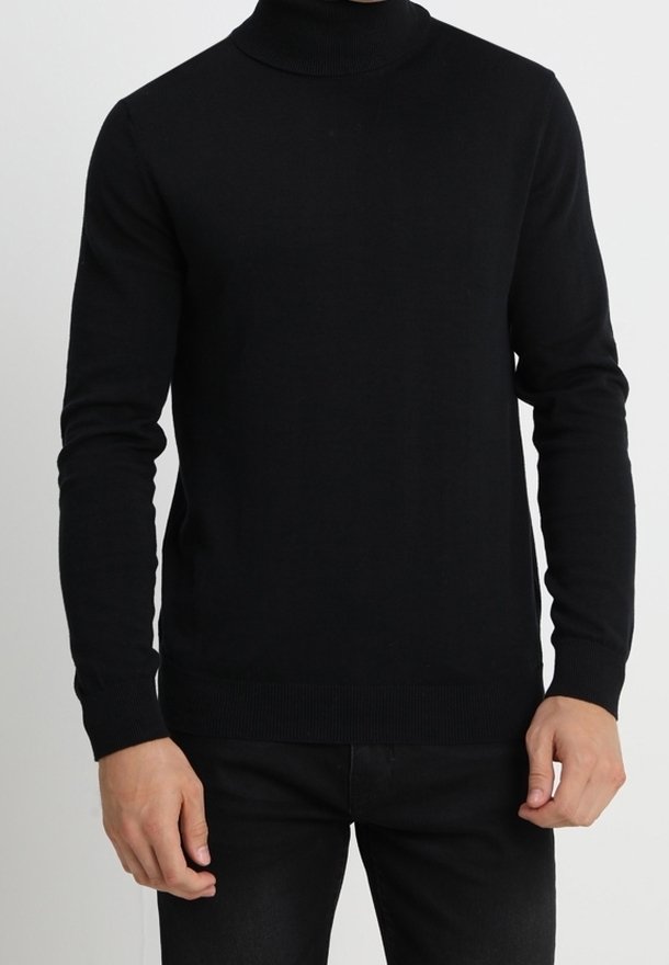 Homme portant un pull noir moulant à col roulé avec manches longues et pantalon noir, debout devant un fond uni.