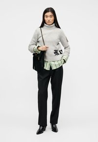 Νεαρή γυναίκα που φοράει γκρι πουλόβερ με ζιβάγκο πάνω από πράσινο ριγέ πουκάμισο, μαύρο παντελόνι, μαύρα μποτάκια αστραγάλου και κρατάει μια μαύρη τσάντα ώμου με φουντίτσες.