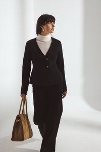 Musternäidisega must bleiser sügava V-kaelusega ja kahe nööbiga, millele on lisatud kreemika värvi kõrge kaelusega särk ja mustad püksid; kannab helepruuni sametist kott.