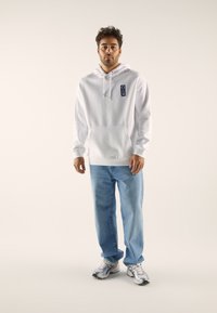 White hoodie with a kangaroo pocket and logo patch, paired with light blue jeans and gray sneakers. Smooth texture throughout.