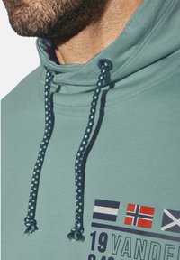 Light teal hoodie features a high collar with braided navy drawstrings and printed flags. Smooth fabric with a functional design.