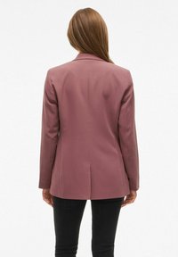 Sako prilagođenog kroja od mekog mauve materijala; ima jedan stražnji prorez, strukturirane ramena i duge rukave. Nošen s crnim hlačama.