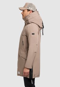 Chaqueta beige resistente a la intemperie con cuello alto, bolsillos con cremallera, capucha ajustable y detalles de abertura lateral, llevada con una gorra con malla en la parte posterior.