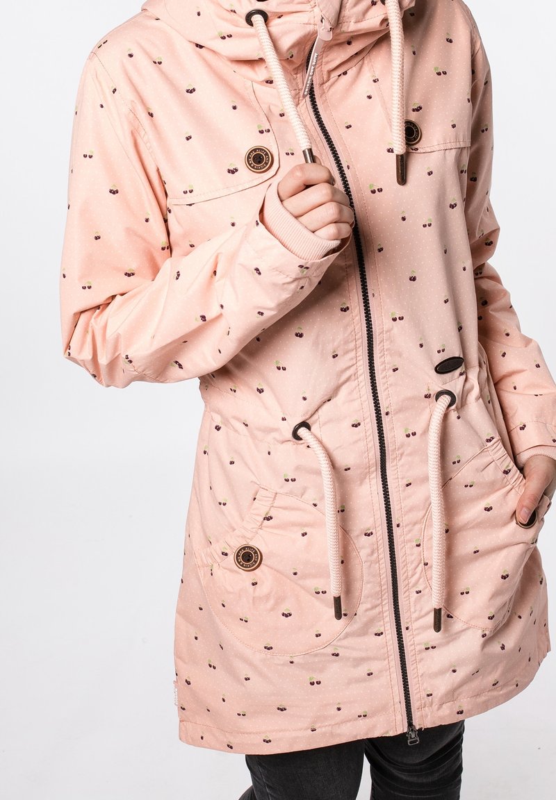 Veste à capuche rose clair avec petits motifs de cerises, fermeture éclair frontale, cordons de serrage, poches à boutons et poignets côtelés sur pantalon noir.