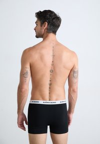 Sorte bomullsboxershorts med en hvit midjebånd som har "BJÖRN BORG" logo. Modellen har intrikate tatoveringer langs ryggraden.