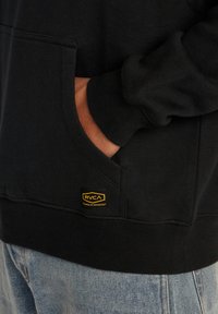 Haut à capuche noir en coton, avec une poche kangourou et des poignets côtelés. Le logo RVCA est affiché en jaune sur le côté inférieur gauche.