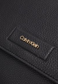 Svart strukturerad läderhandväska med en guld "Calvin Klein"-logotyp på en placerad patch. Designen betonar släta linjer och minimalistisk elegans.