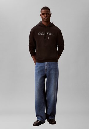 Mann trägt schwarzen Calvin Klein Hoodie, blaue weit geschnittene Jeans und schwarze Schuhe, steht mit den Händen in den Taschen vor einem schlichten grauen Hintergrund.