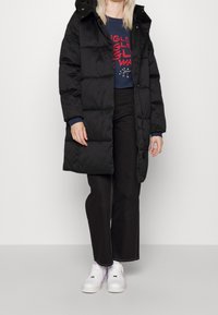 Person som bär en svart puffjacka över en marinblå tröja med röd text, svarta vida byxor och vita sneakers, stående mot en enfärgad bakgrund.