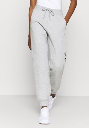 Persoon in lichtgrijze joggingbroek met trekkoord en witte sneakers, lopend met handen in de zakken tegen een effen achtergrond.