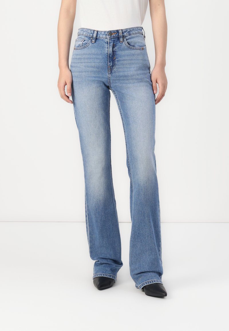 Blaue hochgeschnittene Bootcut-Jeans mit einer glatten Denim-Oberfläche, dezenten Ausbleichungen und einem Fünf-Taschen-Design. Kombiniert mit schwarzen Absatzschuhen.
