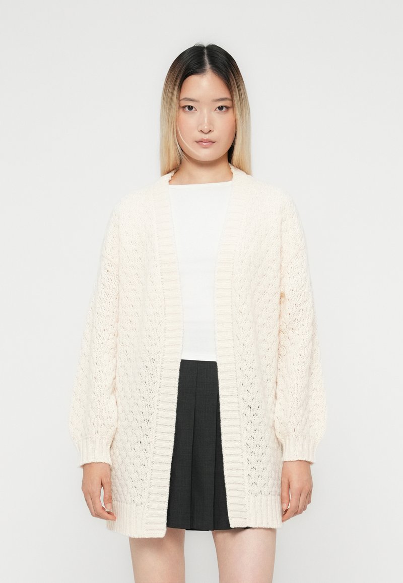 Jeune femme portant un cardigan en tricot crème sur un dessus blanc et une jupe plissée noire, se tenant devant un fond clair et uni.