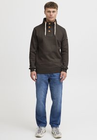 Pullover in pile marrone con colletto alto, tre bottoni e cordini. Abbinato a jeans blu e sneakers chiare.