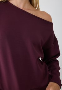 Suéter burdeos con escote off-the-shoulder, en una tela suave y lisa, con mangas largas y un corte holgado. Diseño minimalista con costuras sutiles.