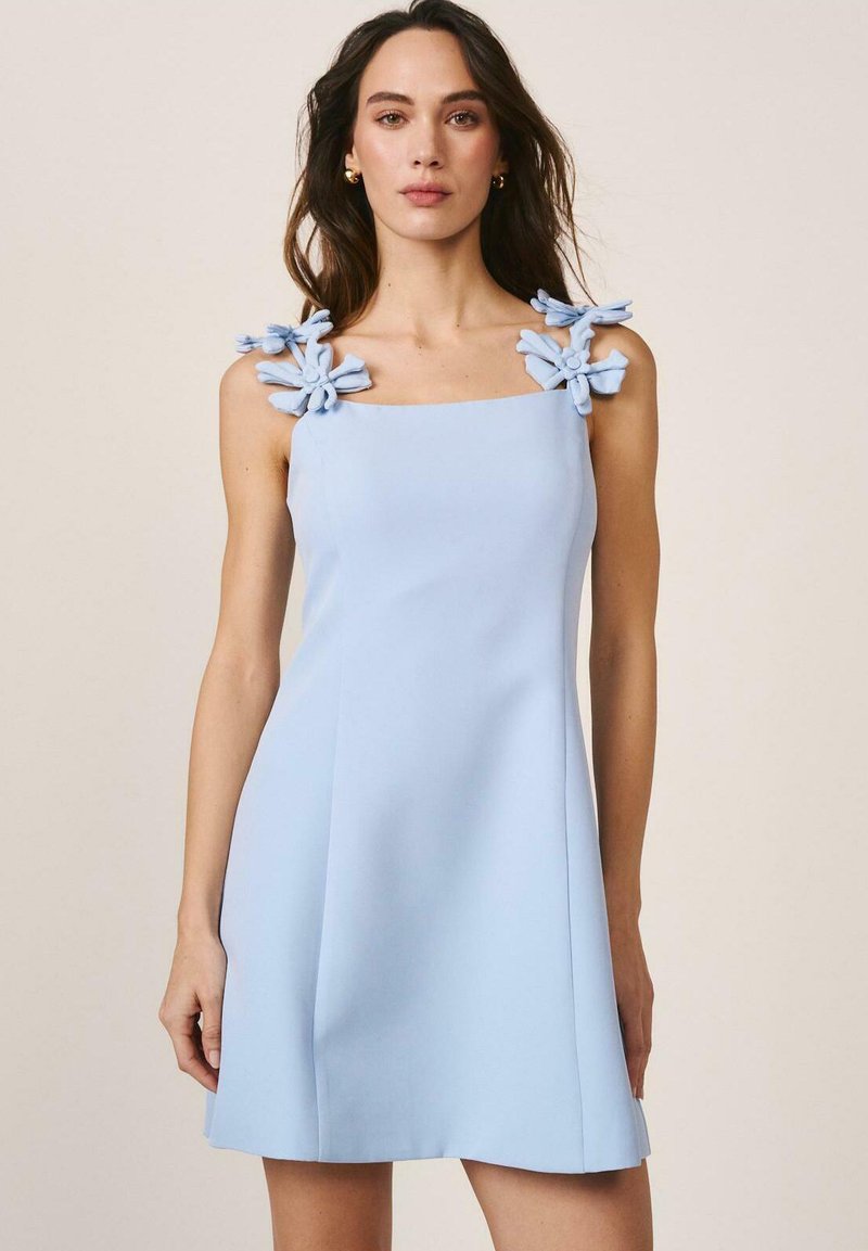 Mujer con un vestido azul claro sin mangas con grandes detalles de flores de tela en las tiras de los hombros, de pie contra un fondo beige liso.