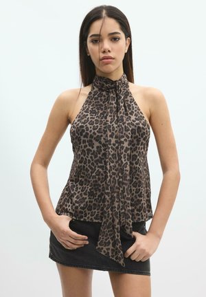 Mladá žena nosí tielko bez rukávov s leopardím vzorom a viazaním a čiernu rifľovú mini sukňu, stojí so založenými rukami vreckách.