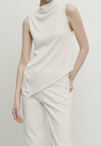 White sleeveless turtleneck top with a diagonal hem, textured knit fabric, paired with white pants, creating a monochromatic look.