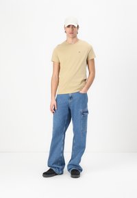 Beige pamut póló, kék laza szabású farmer cargo nadrág oldalsó zsebekkel, fekete cipő és fehér baseball sapka. Minimalista dizájn, laza stílus.