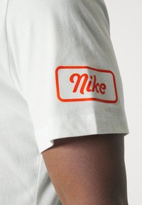 Biała koszulka z krótkimi rękawami z pomarańczowym logo Nike na lewym rękawie. Wykonana z miękkiej bawełny, klasyczny krój z okrągłym dekoltem.