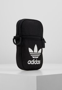 Czarna saszetka Adidas z boczną kieszenią na zamek, białym logo trefoil i tekstem logo, ustawiona pionowo na białej powierzchni na szarym tle.