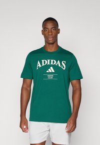 T-shirt de algodão verde apresentando o texto e o logótipo "ADIDAS" em creme, com "TENNIS CLUB" por baixo. Cortes padrão com mangas curtas.