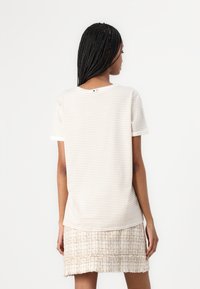 Camiseta de algodón a rayas en crema y beige, con mangas cortas y un cuello redondo. La parte de atrás presenta un pequeño detalle de botón negro.