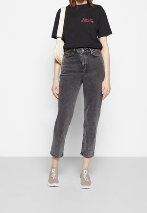 Osoba w czarnej koszulce z czerwonym napisem, w wyblakłych czarnych dżinsach z wysokim stanem, beżowych trampkach i z beżową torebką noszoną na ramieniu.