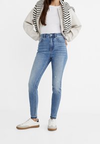 Jeans skinny de tiro alto en azul claro con una textura suave, ligero desgaste y diseño de cinco bolsillos, combinados con una camiseta blanca y zapatillas.