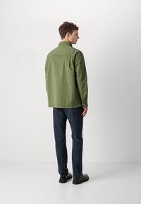 Zelená lehká bunda s límečkem na knoflíky, detailem na zadním díle a dlouhými rukávy, kombinovaná s tmavě modrými džíny a černými botami.