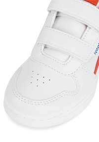 Bijele kožne tenisice s čičak trakama, perforiranim nožnim dijelom i narančastim naglascima sa strane. Elegantan dizajn s šivanim detaljima.