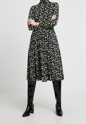 Femme debout, les bras croisés, portant une robe midi noire à motifs floraux blancs et des bottes noires brillantes montant jusqu'aux genoux.