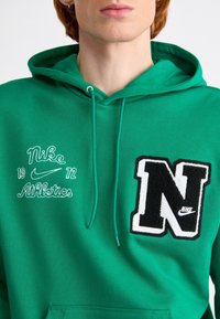 Grön huvtröja med dragsnöre, som har vit broderad "Nike" text, "1972," och svart och vit "N" patch. Mjukt, strukturerat tyg.