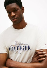 Lys grå bomulds t-shirt med et marineblåt logo med blomsterdesigns. Korte ærmer og rund hals. Glat tekstur.