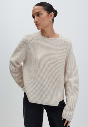 Femme aux cheveux bruns en chignon, portant des boucles d'oreilles en argent, un pull beige à côtes et un pantalon noir, posant contre un fond clair uni.