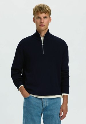 Jeune homme aux cheveux blonds courts portant un pull noir à demi-fermeture sur une chemise blanche et un jean bleu, debout avec une main dans la poche.