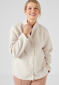 Crème-kleurige fleecejas met een gestructureerd oppervlak, een hoge kraag, voorknopsluiting en contrasterende bies langs de randen. Ontwerp met een ontspannen pasvorm.