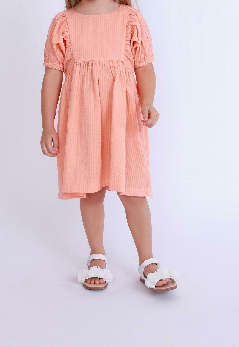 Jeune enfant portant une robe couleur pêche avec des manches bouffantes et des sandales blanches ornées de fleurs, debout devant un fond clair et uni.