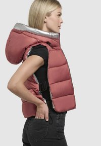 Vesta căptușită în nuanță mauve, cu glugă căptușită cu un fir argintiu strălucitor și închidere cu fermoar. Dispune de catarame laterale și un design ajustat.